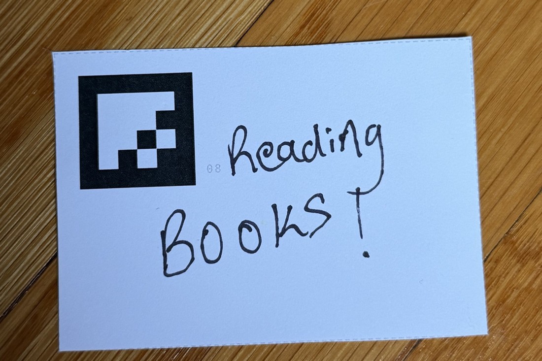 One card on a wooden floor: a printed marker in the corner, and beside it, handwritten, &ldquo;Reading BOOKS!&rdquo;.
